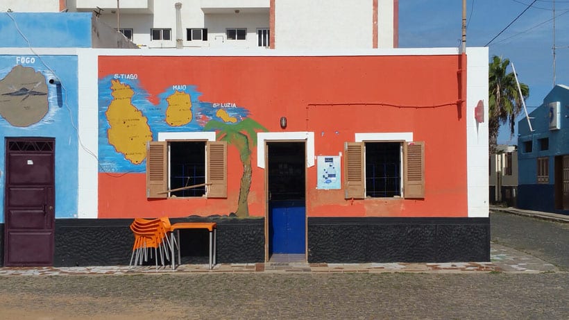 Explore the best things to do in santa maria cape verde, brightly coloured red and blue building with hand painted maps of local islands and a palm tree on the side next to a stack of chairs and a table