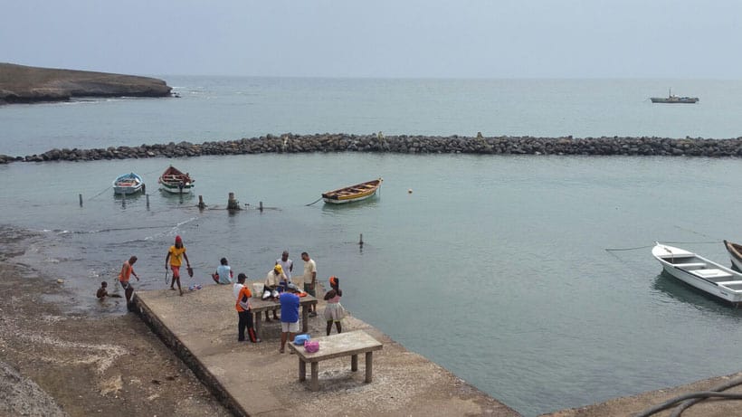 Explore things to do in santa maria cape verde, people standing on a jetty next to wooden tables with boats moored nearby  to a low rock seawall with ocean waters in the distance