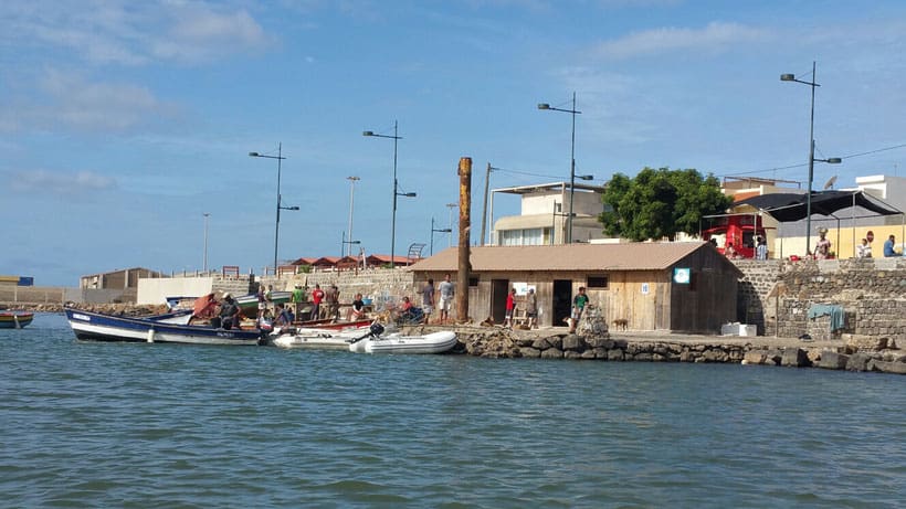 Enjoy shopping in Sal, Cape Verde, boats moored by wooden hut next to wall running along street with people walking by under a blue sky with clouds