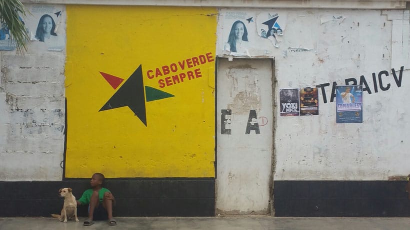 Find the best things to do in santa maria, cape verde, small child sitting with dog in front of building with colourful yellow painted wall featuring black star with red and green points and the words "Cabo Verde Sempre"