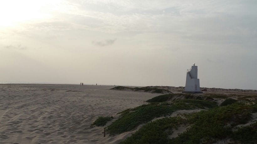 Wander around new places to visit in sal cape verde, white sandy beach with areas of green foliage next to tall viewing point structure and people in the distance