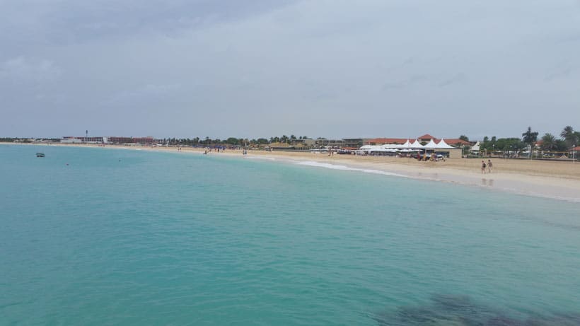 Enjoy the best things to do in sal cape verde, clear turquoise waters lapping gently at a long sandy beach with people walking along the shore next to small buildings and huts