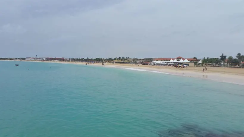 Enjoy the best things to do in sal cape verde, clear turquoise waters lapping gently at a long sandy beach with people walking along the shore next to small buildings and huts