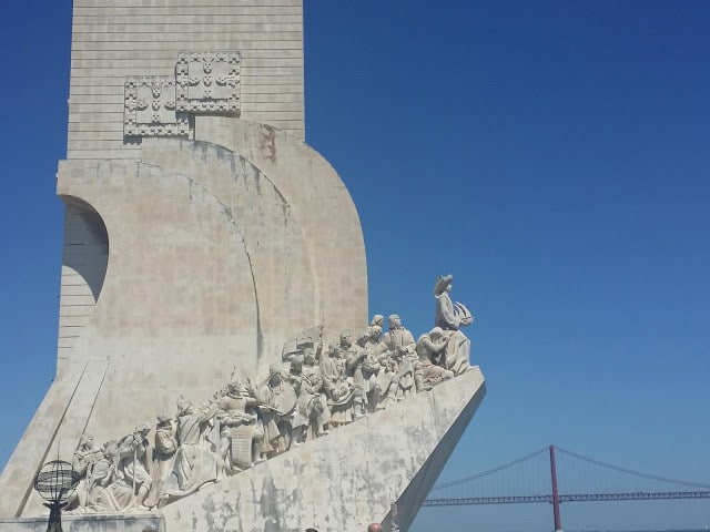 a closeup of a statue with people climbing upwards with a bridge in the background of lisbon