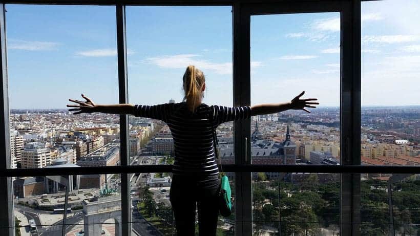  A woman stands with her back to the camera, silhouetted against a window overlooking the cityscape of Madrid. Her arms are outstretched widely. She is wearing a black jacket and black leggings, and has blonde hair. The window is wide, and the city view shows many buildings, streets, and trees. 