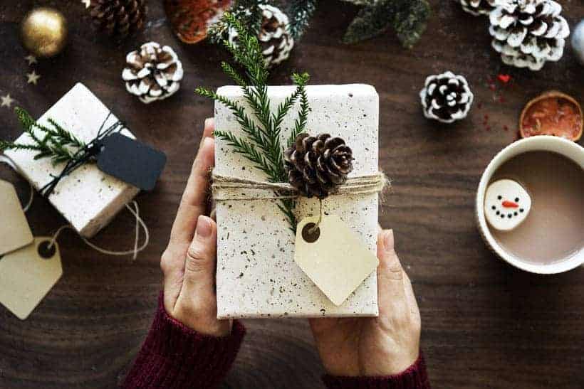 person holding Christmas gift wrapped in handmade paper with pinecone and tree sprig, on table below is a similarly wrapped gift, more pinecomes and hot cocoa with snowman marshmallow