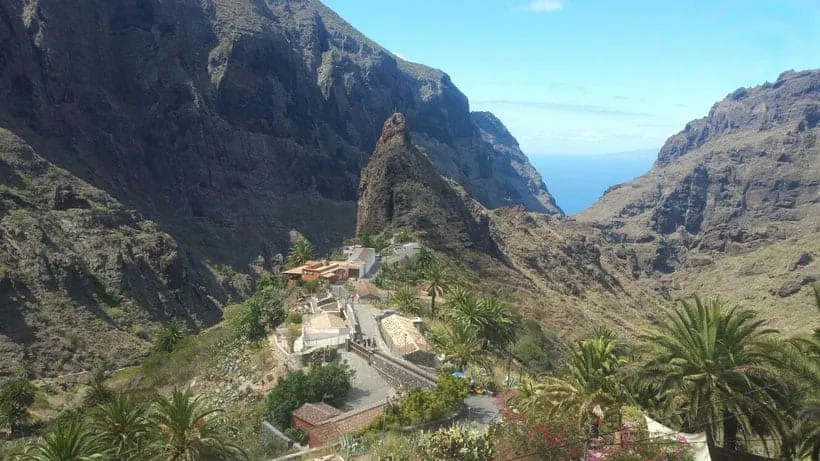 Discover the best walks in Tenerife, view of Masca Gorge with large rocky cliffs and central outcrop with road leading up to it and blue sea in the distance