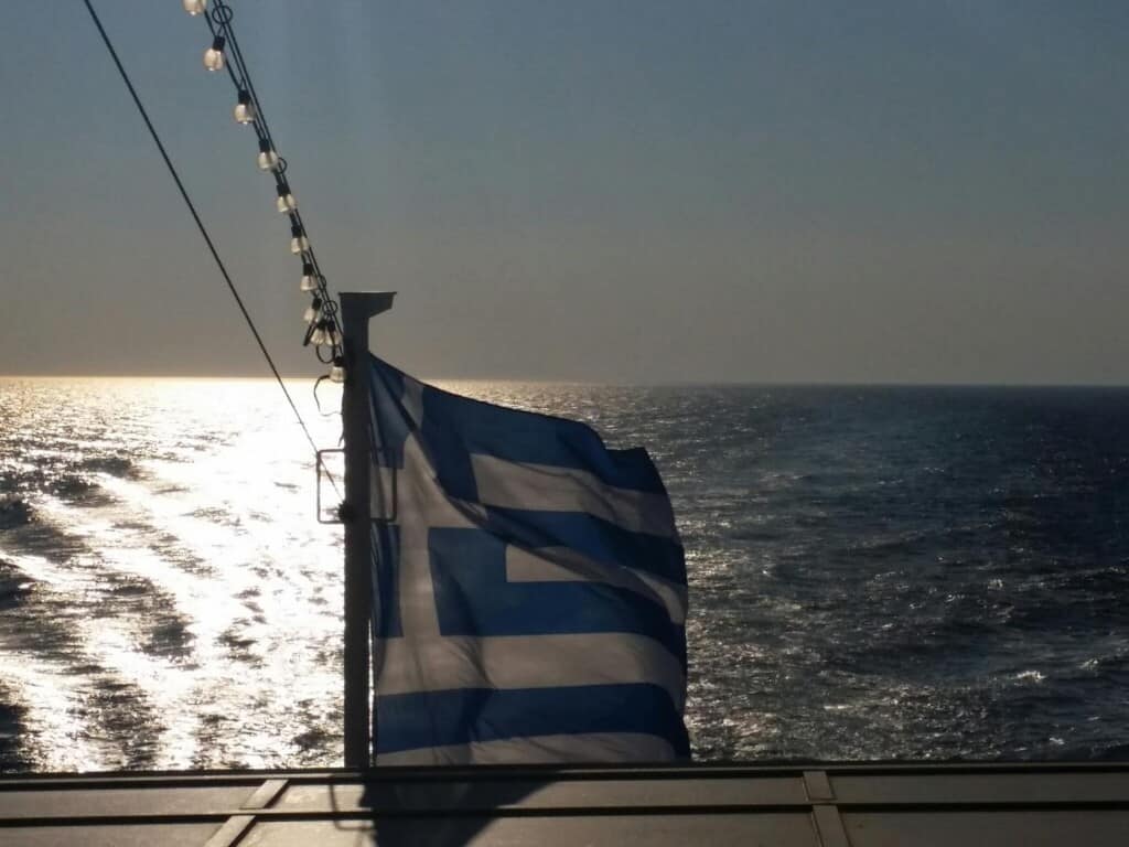 A Greek flag waves in the wind on a ferry with the sun setting in the background. 