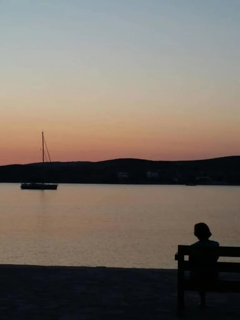 must see things in paros, person on bench watching boat in the sea at sunset