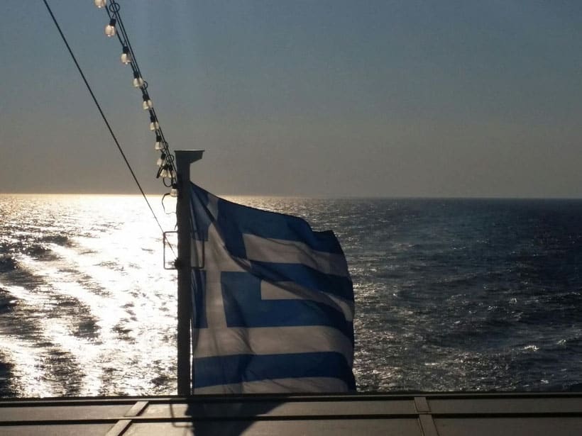 why you should visit paros greece, greek flag blowing in wind over the sea