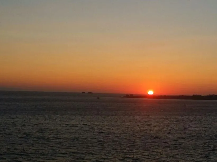 A breathtaking sunset paints the sky with vibrant hues of orange and pink, casting a warm glow on the calm Aegean Sea in Paros, Greece. Silhouettes of distant islands dot the horizon, adding to the tranquil beauty of the scene.