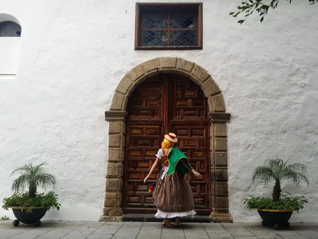Best places to visit in Tenerife, person wearing traditional costume standing in front of large ornate wooden door in arched stone doorway set in a white walled building with green potted plants to either side