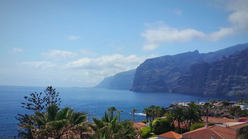 Why you should move to Tenerife, view of large rocky cliffs on coastline next to deep blue sea under a cloudy blue sky with palm trees and terracotta rooftops in the foreground