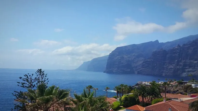 Why you should move to Tenerife, view of large rocky cliffs on coastline next to deep blue sea under a cloudy blue sky with palm trees and terracotta rooftops in the foreground