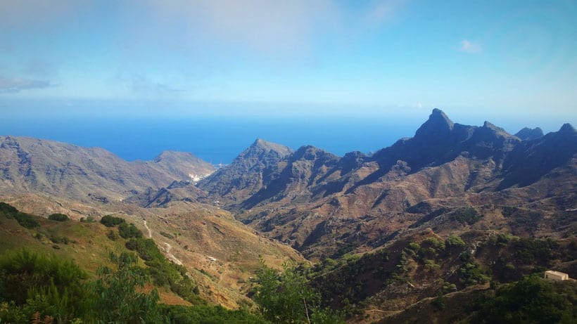 Ways to start living in Tenerife, view of valley with large green rocky mountains and grass covered hills leading down towards blue sea under an azure blue sky
