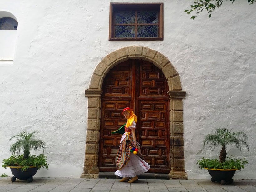 Best Reasons to Move to Tenerife, person in traditional local dress standing in front of large wooden double door set into white stone wall of building with grey archway and potted plants either side
