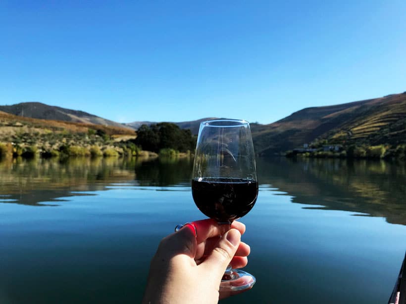 things to do in douro valley portugal, person holding up wine glass with red wine overlooking a large body of water on clear a sunny day with mountains in the distance