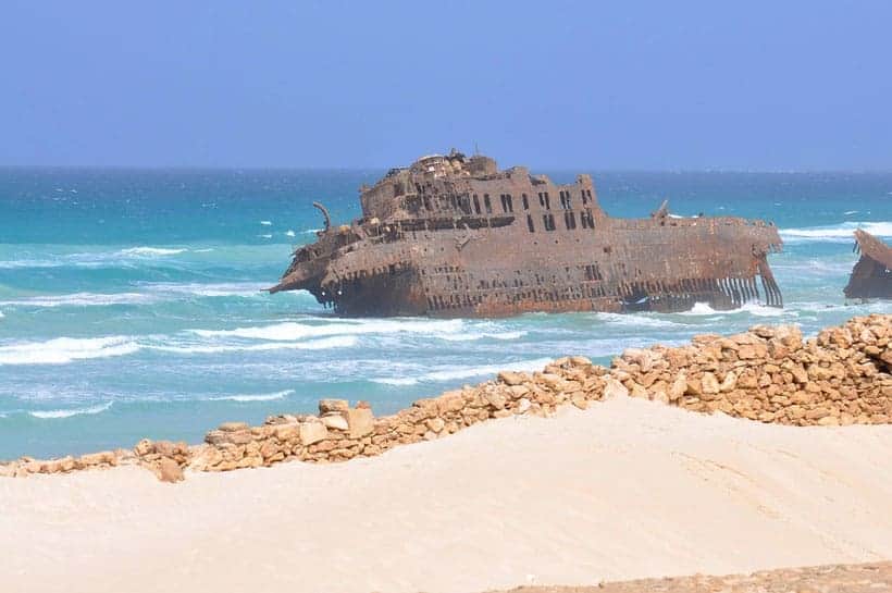 rusted remains of shipwreck sitting in the waves just offshore with white sand and a low rock wall in front and the vast blue expanse of the sky behind