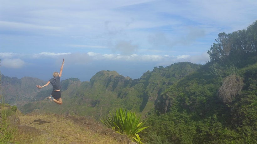 a woman jumping on a view point with the view of the sky, mountains, and clouds