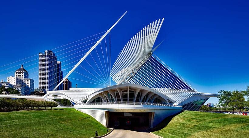 unique things to do in Wisconsin in January, view of Milwaukee Art Museum with modern architecture comprised of many white beams and cable supports flanked by two steep verges of green grass under a clear azure blue sky