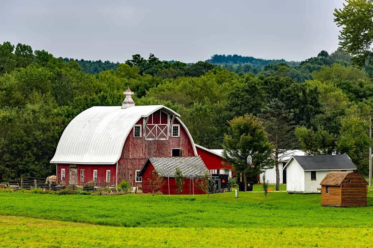 Places to visit in Wisconsin in March, Large red and white barn with accompanying smaller sheds surrounded by large fields of green grass and green trees all under a bright grey sky