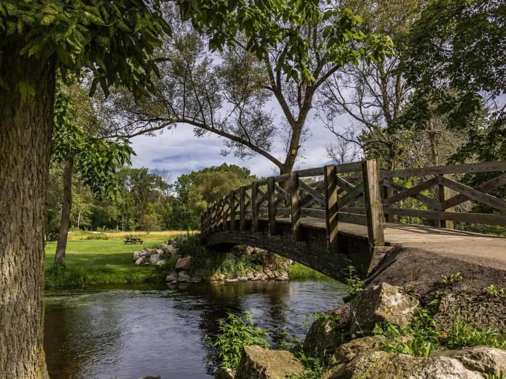 best excursions from Milwaukee, wooden bridge over a river in Cedarburg, WI with green grass and trees beyond under a bright cloudy sky