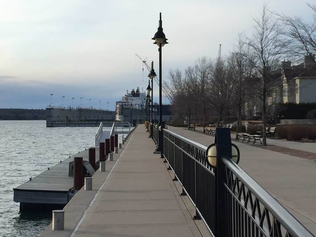 a lakeside boardwalk by a small dock, with a street with trees with no leaves