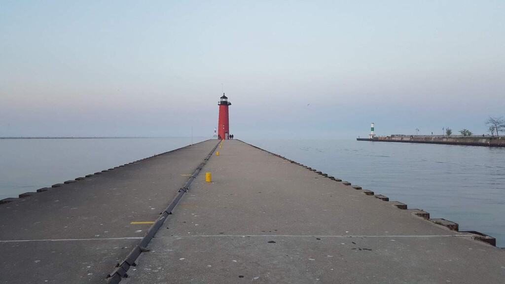 Enjoy some of the best fall activities near Milwaukee, view looking along a pier towards a tall red lighthouse standing on the edge of a lake under a darkening sky