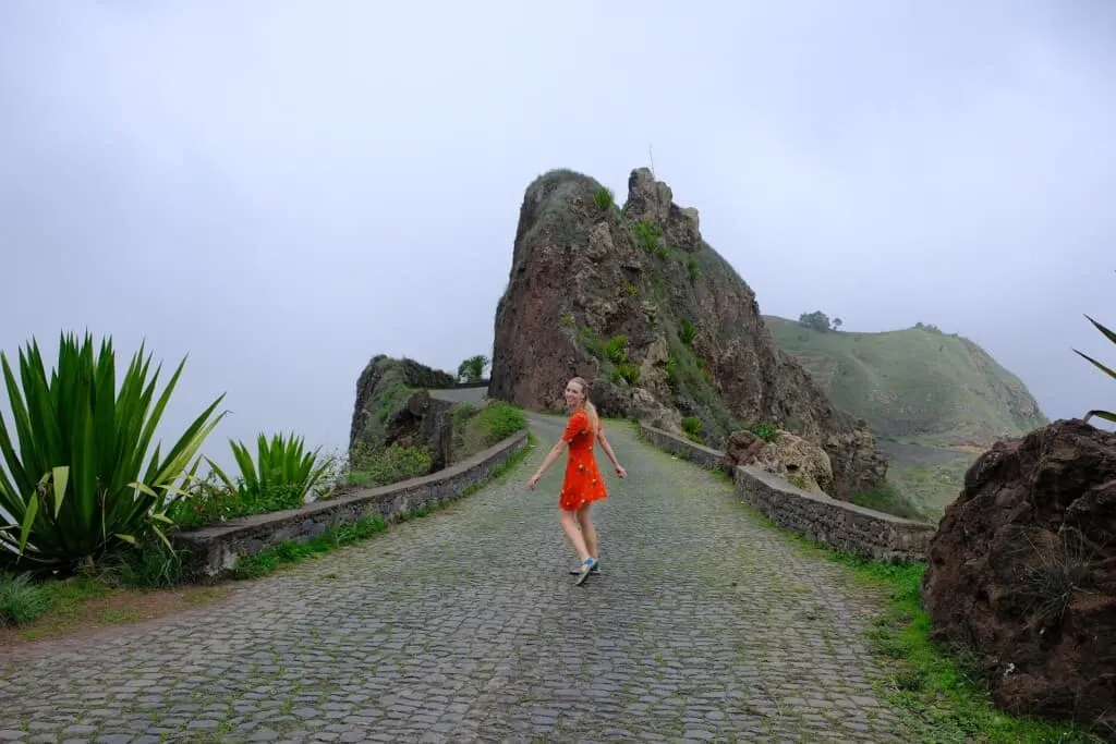 delgadim santo antao, a woman in red looking at the camera but walking towards a pathway on a cloudy day