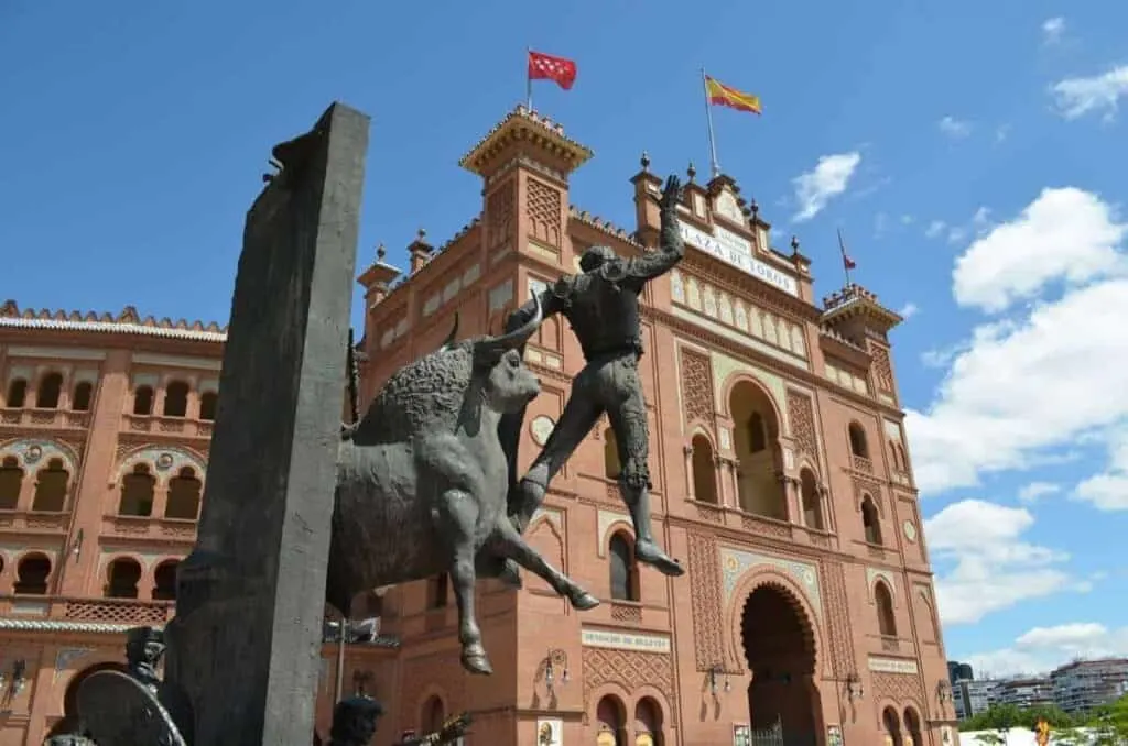 The Bullfighting Museum in Madrid stands against a blue sky. It is a sizeable Spanish-style building with intricate windows made of terracotta-colored stone. In the foreground of the museum is a giant iron statue of a bull with a bullfighter standing in front. 