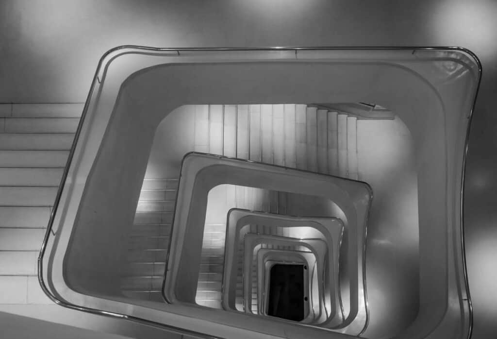 A white cubical staircase is photographed from above, creating the illusion of a deep tunnel. The staircase is located in CaixaForum in Madrid, Spain. 