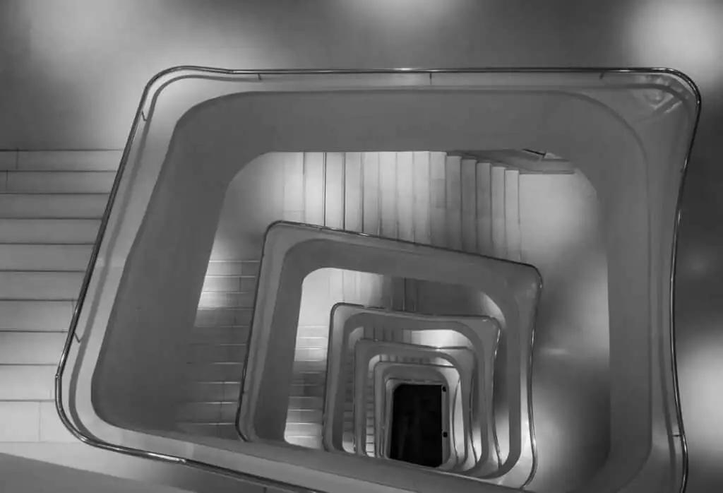 A white cubical staircase is photographed from above, creating the illusion of a deep tunnel. The staircase is located in CaixaForum in Madrid, Spain. 