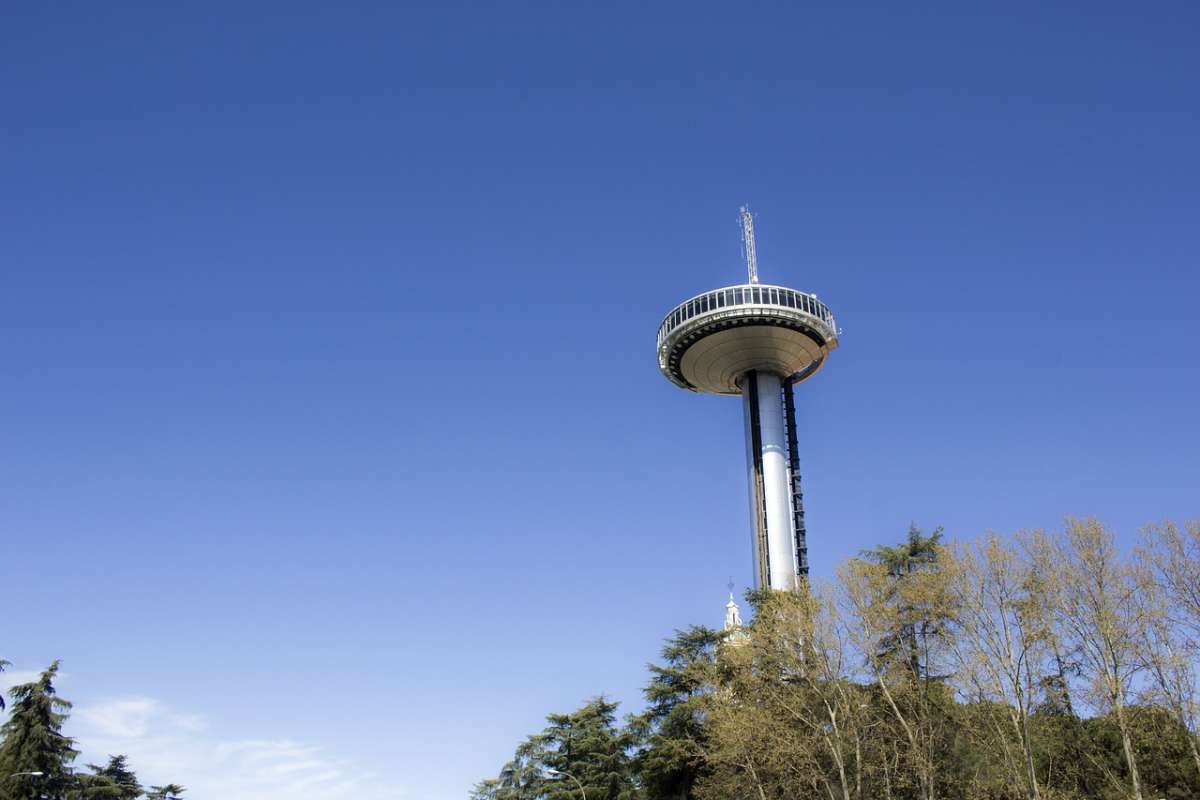 The Faro de Moncloa stands against a blue sky in Madrid, Spain. The Structure is a tall tower with a round platform on top, featuring windows. Visitors can look out from the platform and see the trees and city below.