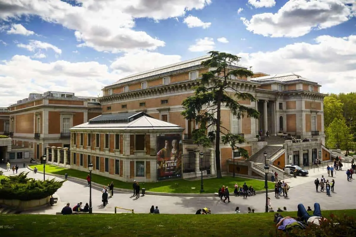 A landscape image of the castle-like El Prado Museum in Madrid, Spain on a clear, sunny day against a blue sky. People walk along the paths and relax in the grass in front of the museum. 