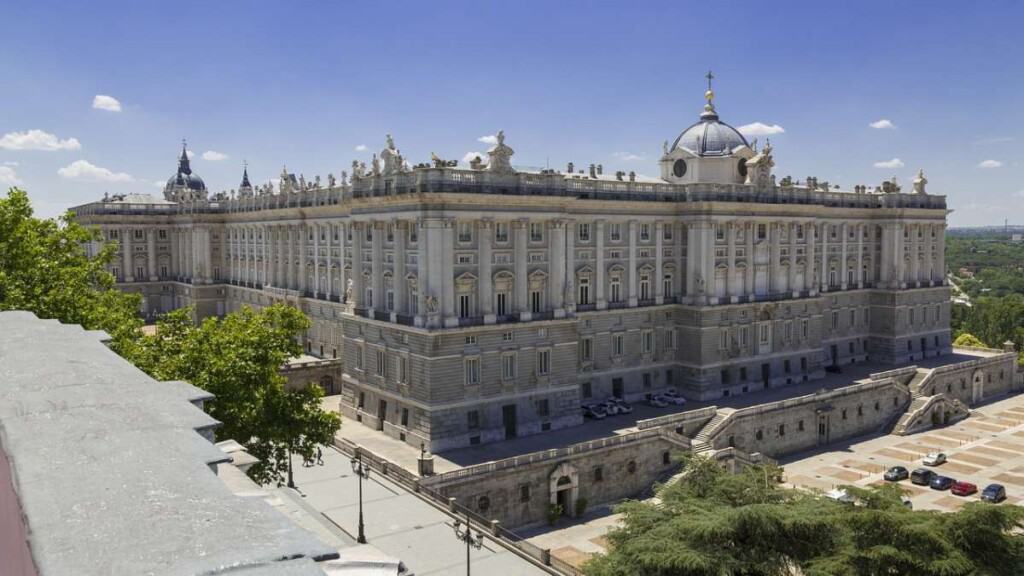 The Royal Palace of Spain is a huge light-colored stone structure covering a square city block. In this image, the Palace sits against a blue sky, along a city street surrounded by trees. 