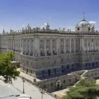 The Royal Palace of Spain is a huge light-colored stone structure covering a square city block. In this image, the Palace sits against a blue sky, along a city street surrounded by trees.