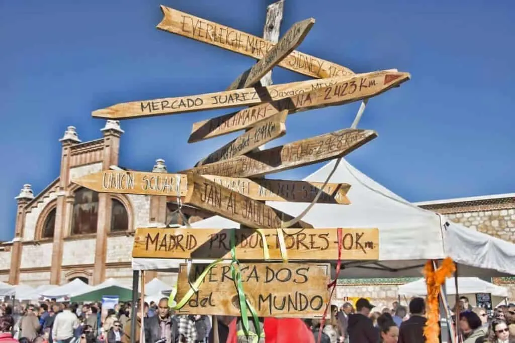 A wooden fingerpost sign stands in the foreground of a bustling marketplace. Tents are set up in the background, and a large crowd explores the El Matedero market. There is a Spanish-style stone building with four turrets against a bright blue sky in the back. 