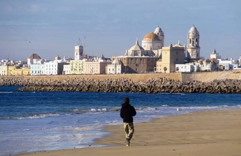 Where to go in Spain in February a person walking on a white sand beach near the waves with a view of a town with white painted buildings and a dome church