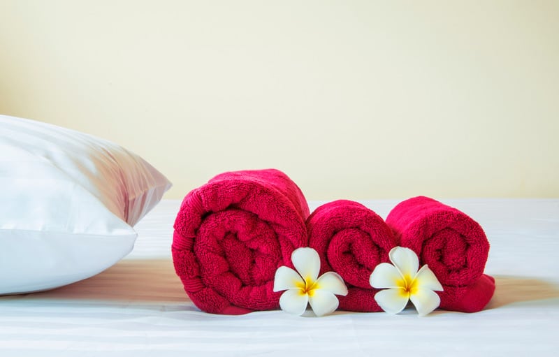 close up shot of three red towels neatly folded and placed on top of a white bed next to a soft white pillow with two delicate and decorative white and yellow flowers