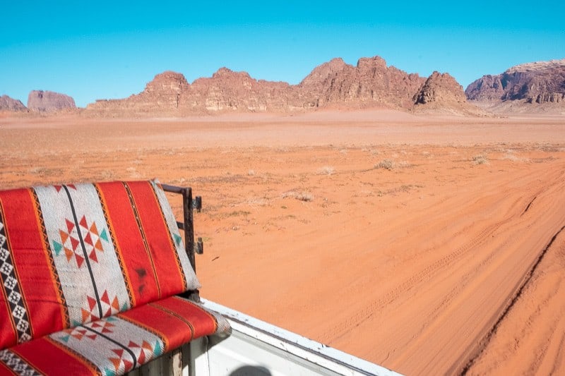 Discover the best free things to do in Wadi Rum, view from the back of a truck driving through the desert with rocky mountains behind under a bright blue sky