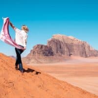 What can you do in Wadi Rum this summer, person walking up a sand dune while holding a patterned shawl with large rocky mountains in the distance under a clear blue sky
