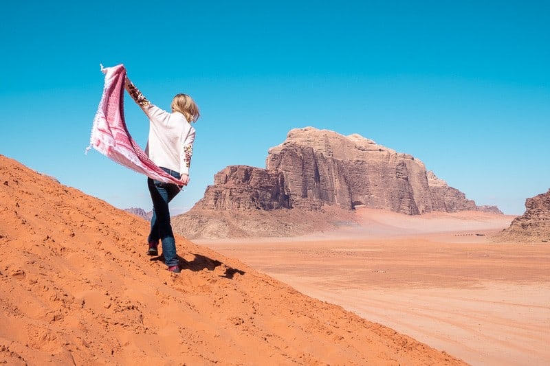 What can you do in Wadi Rum this summer, person walking up a sand dune while holding a patterned shawl with large rocky mountains in the distance under a clear blue sky