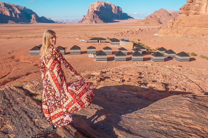 Find some beautiful places in Wadi Rum, person in long patterned dress standing in front of view of the Best Bedouin Camp in Wadi Rum with large rocky mountains in the distance under a bright blue sky