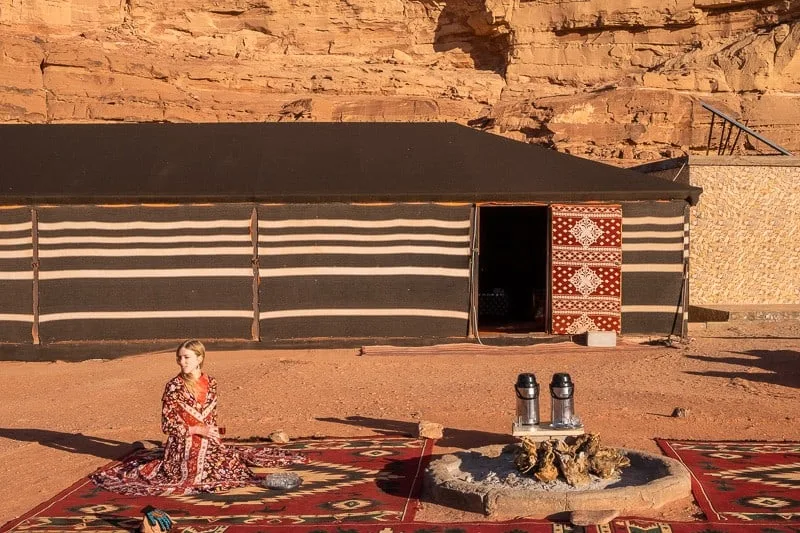 Find the best places to see Wadi Rum, person sitting on large ornate rug next to fire pit outside Bedouin camp dwelling in the sun