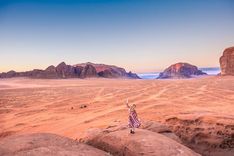 Plan the perfect Wadi Rum itinerary, person standing on large rocks in front of wide view of the desert under a clear sky at sunset