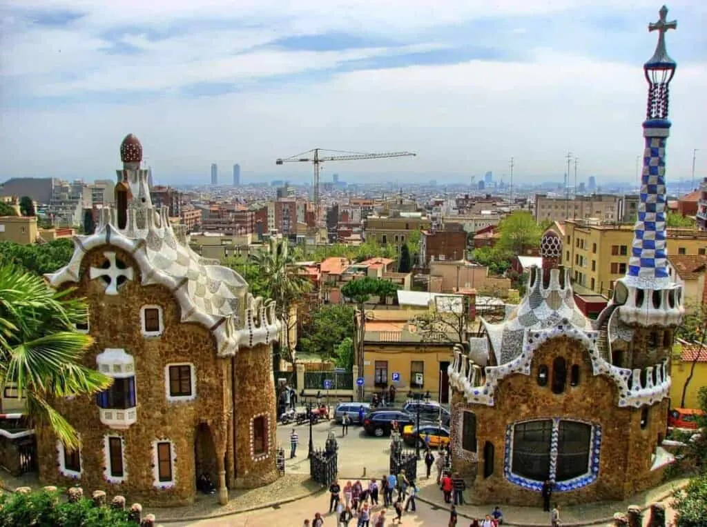 two buildings in parc guell with a tower at the back, other buildings and people walking around