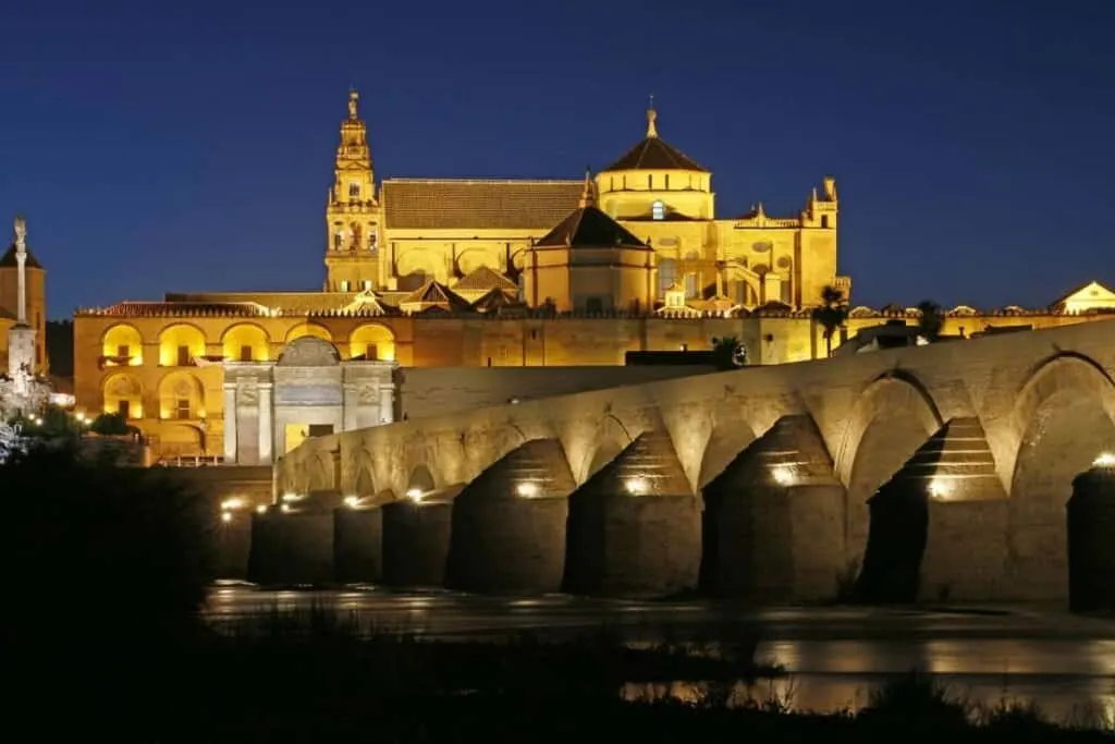 Stay in these amazing spanish paradores, view of the Parador de Cordoba illuminated by yellow spotlights and streetlights at night as it sits on a small hill in front of a stone bridge stretching across a river