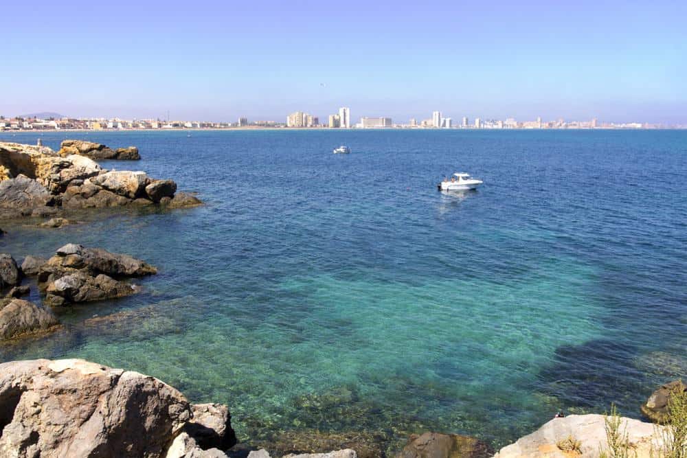 the sea with corals underneath with rocks on the side and incoming boats, a line of buildings on the background