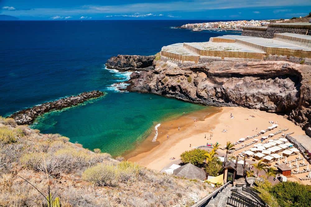 best beaches in tenerife, View down into sandy beach area with sun loungers and awnings on golden sand between large rocky hillsides with green sea water leading out towards dark blue open sea water and built up areas in the distance