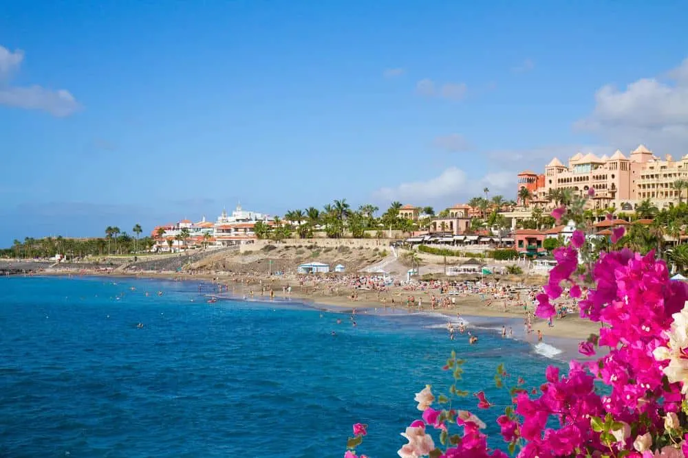 Enjoy the best Tenerife all inclusive adults only hotels, view of sandy beach with people playing in the sun in front of a hotel complex with green trees all under a bright blue sky